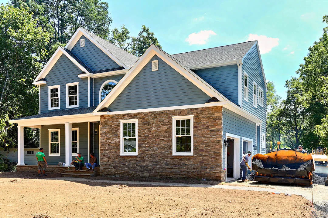 Newly built two-story house with blue siding, a stone front and a covered porch, with crew finishing the driveway.
